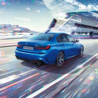 Imagine cu partea laterală spate a BMW Seria 3 Sedan Imagine cu trei sferturi din partea spate a BMW Seria 3 Sedan, rulând pe timpul zilei, în faţa unui peisaj urban