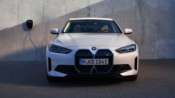 Imagine din partea frontală a unui BMW i4 eDrive40 G26 2021 şi a unui sistem BMW Wallbox Imagine din partea frontală a unui BMW i4 eDrive40 G26 2021 Alb Mineral parcată lângă peretele unei case pe care este montată o staţie de încărcare BMW Wallbox