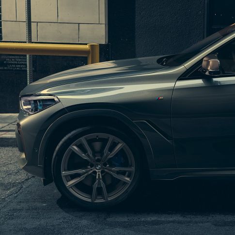 Perspectivă laterală a BMW X6 M50i. Imagine cu un bărbat sprijinit de partea laterală a BMW X6 M50i, pe un fundal urban.