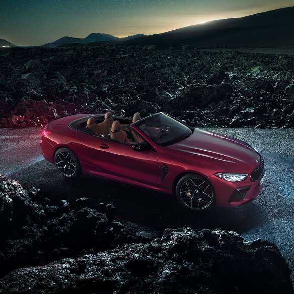 Imagine cu trei sferturi din partea laterală a BMW M8 Competition Cabriolet. Imagine cu trei sferturi din partea laterală a BMW M8 Competition Cabriolet în Roşu Motegi metalizat cu soft-top-ul deschis, staţionând noaptea pe asfalt.