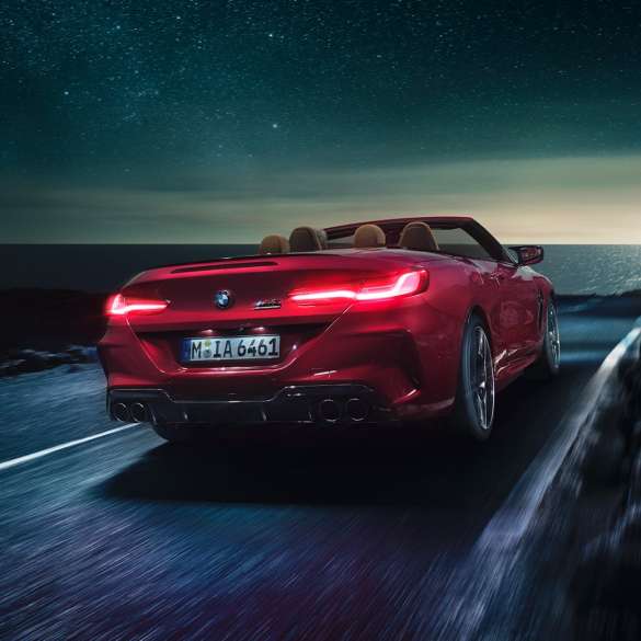 Imagine cu trei sferturi din partea spate a BMW M8 Competition Cabriolet. Imagine cu trei sferturi din partea spate a BMW M8 Competition Cabriolet în Roşu Motegi metalizat cu soft-top-ul deschis, rulând pe timp de noapte de-a lungul coastei.