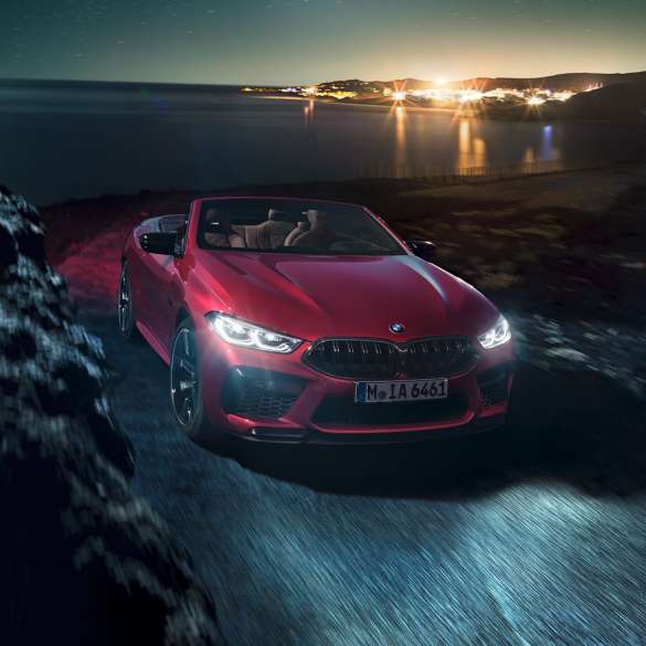 Imagine cu trei sferturi din partea frontală a BMW M8 Competition Cabriolet. Imagine cu trei sferturi din partea frontală a BMW M8 Competition Cabriolet în Roşu Motegi metalizat cu soft-top-ul deschis, rulând pe timp de noapte de-alungul coastei.