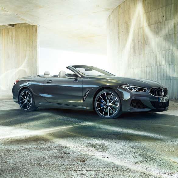 Imagine cu trei sferturi din partea laterală a BMW M850i xDrive Cabriolet. Imagine cu trei sferturi din partea laterală a BMW M850i xDrive Cabriolet în Gri Dravit metalizat cu soft-top-ul deschis, staţionând în faţa unei clădiri de beton.