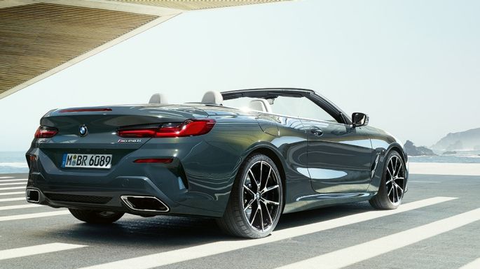 Imagine cu trei sferturi partea spate a BMW M850i xDrive Cabriolet. Imagine cu trei sferturi din partea spate a BMW M850i xDrive Cabriolet în Gri Dravit metalizat cu soft-top-ul deschis, parcând cu oceanul pe fundal.
