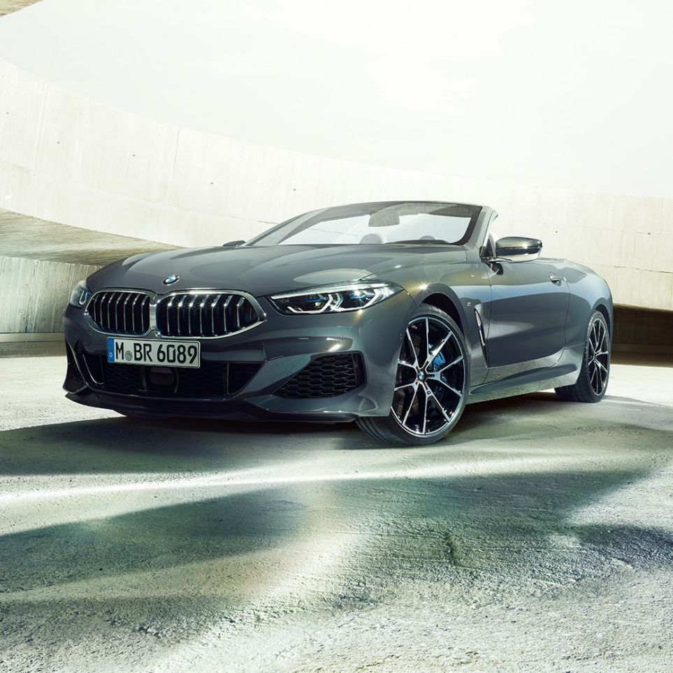 Imagine cu trei sferturi din partea frontală a BMW M850i xDrive Cabriolet. Imagine cu trei sferturi din partea frontală a BMW M850i xDrive Cabriolet în Gri Dravit metalizat cu soft-top-ul deschis, staţionând în faţa unei clădiri de beton.
