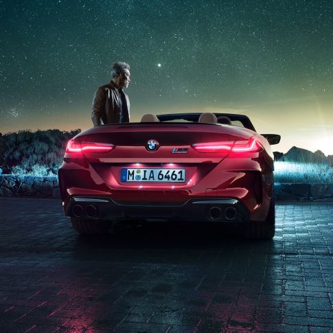 Imagine cu partea spate a BMW M8 Competition Cabriolet. Imagine cu partea spate a BMW M8 Competition Cabriolet în Roşu Motegi metalizat cu capota deschisă, staţionând alături de un bărbat sub cerul înstelat.
