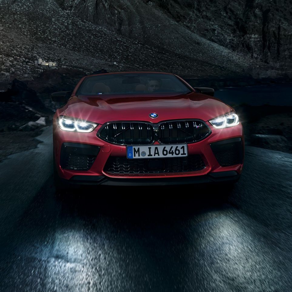 Imagine cu partea frontală a BMW M8 Competition Cabriolet. Imagine cu partea frontală a BMW M8 Competition Cabriolet în Roşu Motegi metalizat, cu faruri strălucitoare, rulând noaptea.