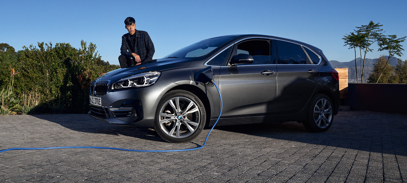 Imagine cu trei sferturi din partea laterală a unui BMW 225xe Active Tourer F45 plug-in hybrid aflat la încărcat Imagine cu trei sferturi din partea laterală a unui BMW 225xe Active Tourer F45 plug-in hybrid aflat la încărcat, cu un model