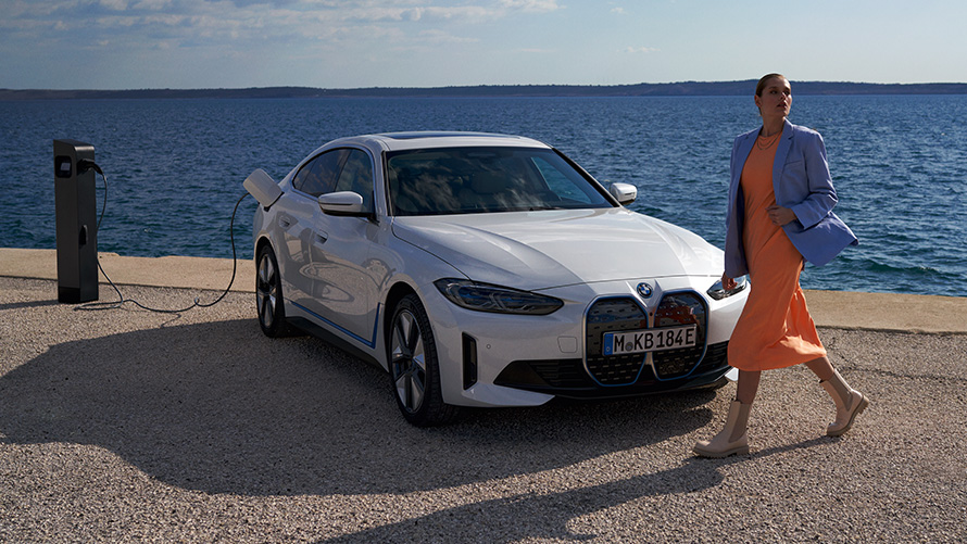 Imagine cu trei sferturi din partea frontală a unui BMW i4 eDrive40 G26 2021 care se află la o staţie de încărcare Imagine cu trei sferturi din partea frontală a unui BMW i4 eDrive40 G26 2021 Alb Mineral care se află la o staţie de încărcare lângă un lac, iar o femeie stă în faţa lui