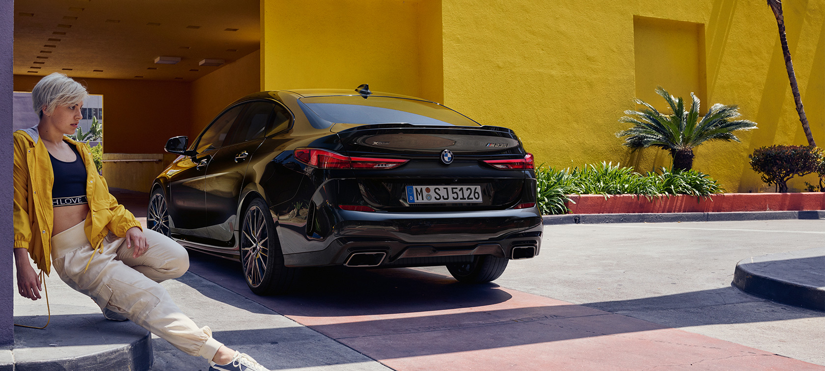 Imagine cu trei sferturi din partea spate a BMW M235i xDrive Gran Coupé Imagine cu trei sferturi din partea spate a BMW M235i xDrive Gran Coupé, în Negru Sapphire metalizat, F44, cu o femeie în faţă