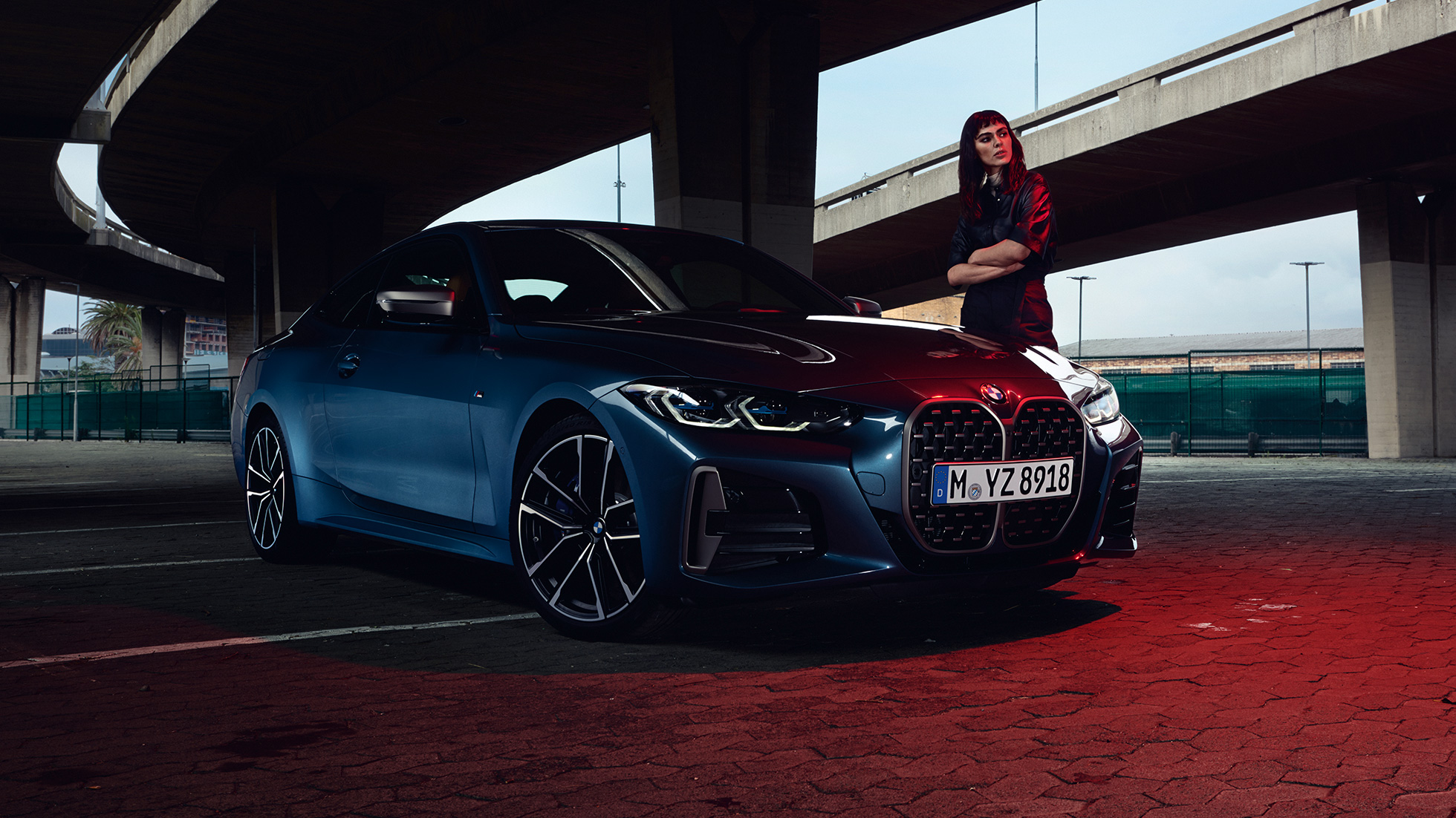 Imagine cu trei sferturi din partea laterală a BMW M440i xDrive Coupé, G22, 2020, cu un model stând lângă automobil Imagine cu trei sferturi din partea laterală a BMW M440i xDrive Coupé, G22, 2020, în Albastru Arctic Race metalizat, cu un model