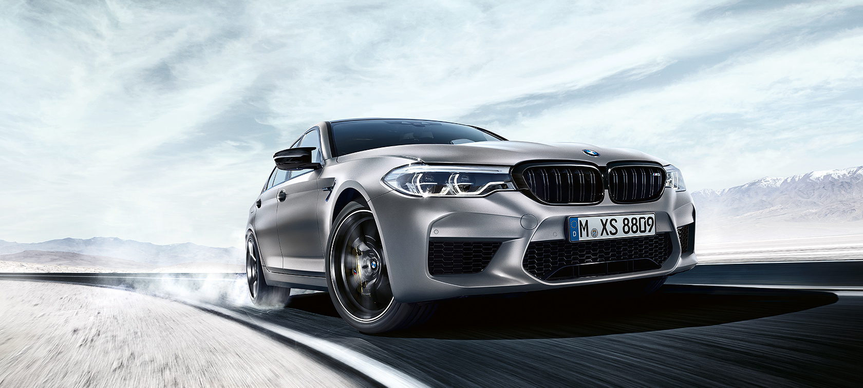 BMW M5 Competition în acţiune BMW M5 Competition gri conduce pe şosea în faţa unui peisaj deşertic