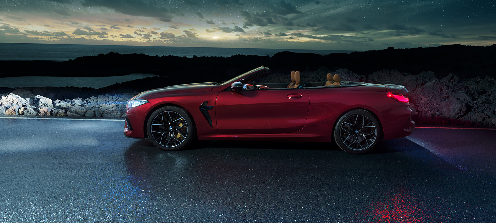 Imagine cu partea laterală a BMW M8 Competition Cabriolet. Imagine cu partea laterală a BMW M8 Competition Cabriolet în Roşu Motegi metalizat cu soft-top-ul deschis, staţionând pe un drum de coastă sub cerul înstelat.