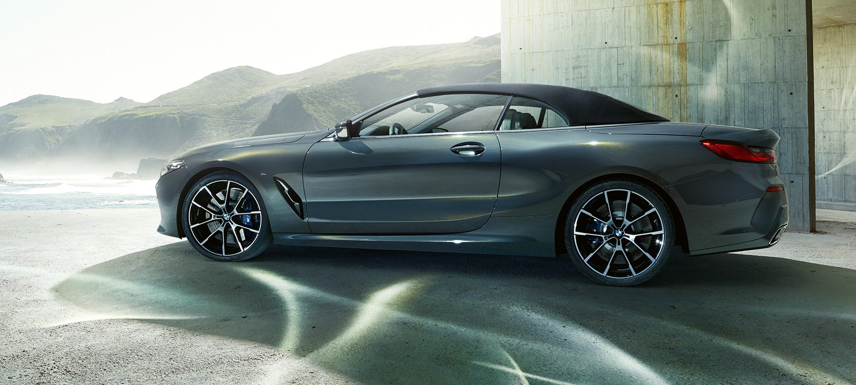 Imagine cu partea laterală a BMW M850i xDrive Cabriolet. Imagine cu partea laterală a BMW M850i xDrive Cabriolet în Gri Dravit metalizat cu soft-top-ul închis, în faţa dealurilor.