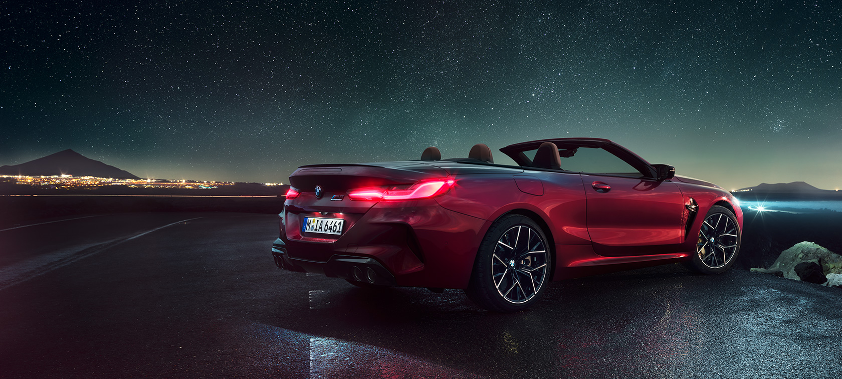 Imagine cu trei sferturi din partea spate a BMW M8 Competition Cabriolet. Imagine cu trei sferturi din partea spate a BMW M8 Competition Cabriolet în Roşu Motegi metalizat cu soft-top-ul deschis, staţionând sub cerul înstelat.