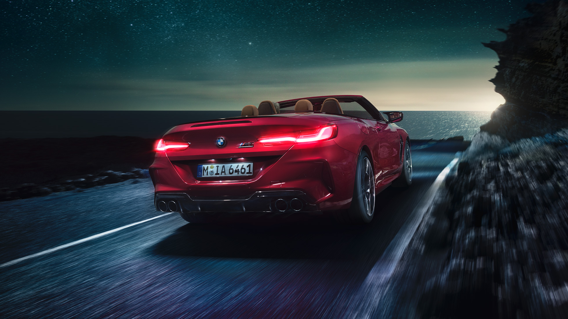 Imagine cu trei sferturi din partea spate a BMW M8 Competition Cabriolet. Imagine cu trei sferturi din partea spate a BMW M8 Competition Cabriolet în Roşu Motegi metalizat cu soft-top-ul deschis, rulând pe timp de noapte de-a lungul coastei.