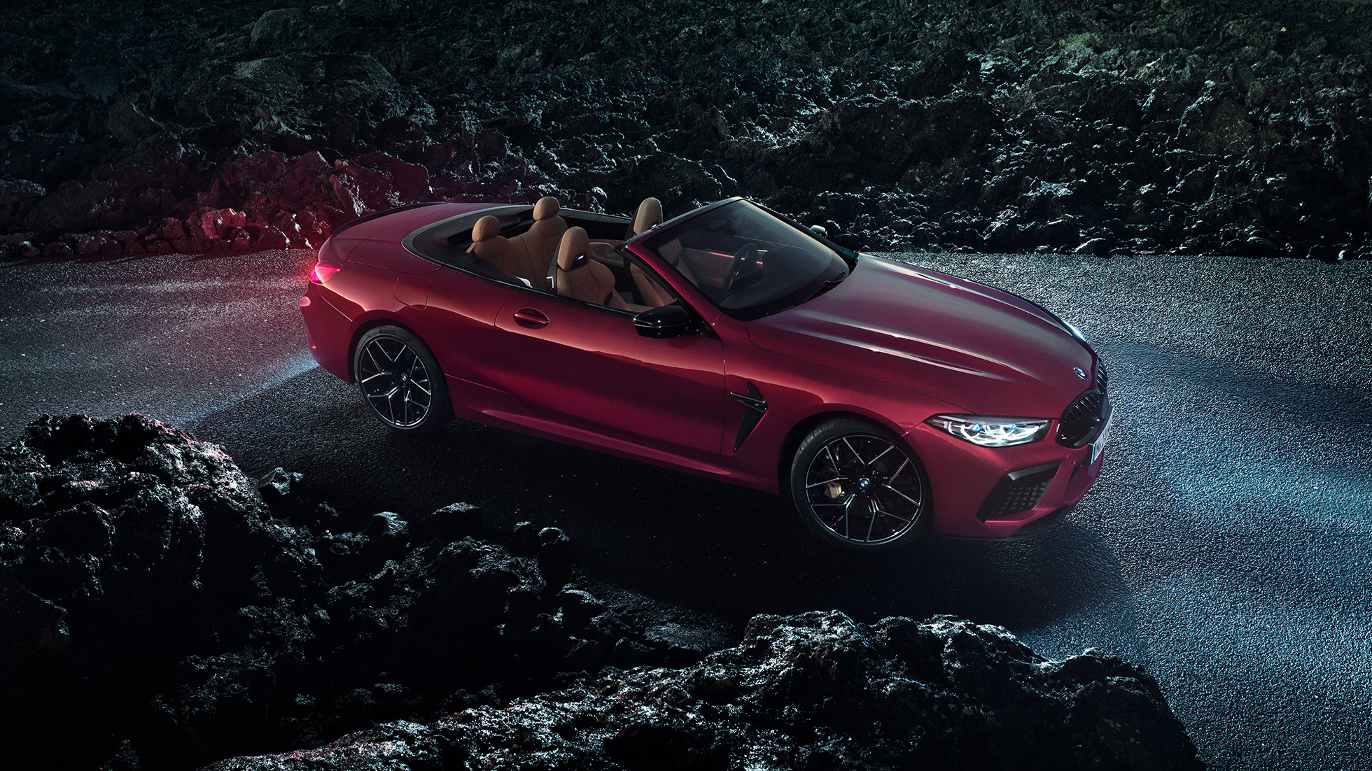 Imagine cu trei sferturi din partea laterală a BMW M8 Competition Cabriolet. Imagine cu trei sferturi din partea laterală a BMW M8 Competition Cabriolet în Roşu Motegi metalizat cu soft-top-ul deschis, staţionând noaptea pe asfalt.