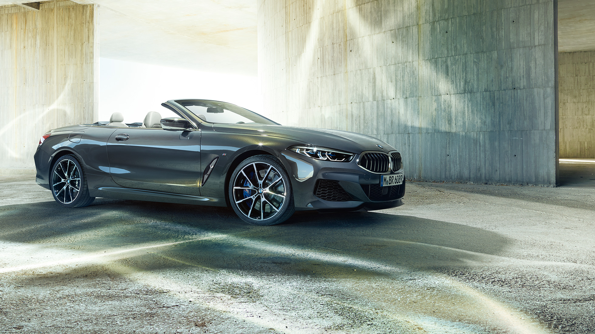 Imagine cu trei sferturi din partea laterală a BMW M850i xDrive Cabriolet. Imagine cu trei sferturi din partea laterală a BMW M850i xDrive Cabriolet în Gri Dravit metalizat cu soft-top-ul deschis, staţionând în faţa unei clădiri de beton.