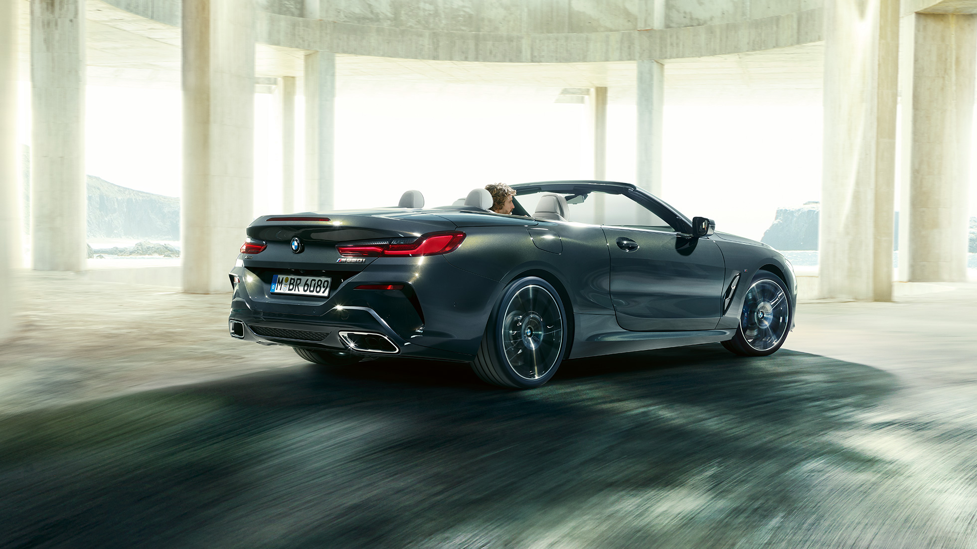 Imagine cu trei sferturi din partea spate a BMW M850i xDrive Cabriolet. Imagine cu trei sferturi din partea spate a BMW M850i xDrive Cabriolet în Gri Dravit metalizat cu soft-top-ul deschis, rulând in interiorul unei clădiri din beton.