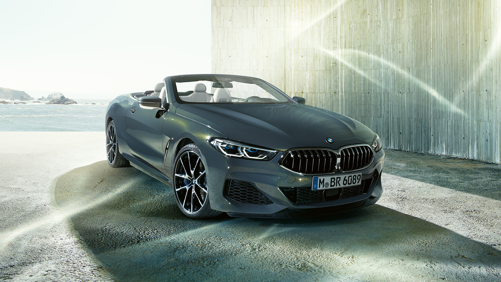 Imagine cu trei sferturi din partea frontală a BMW M850i xDrive Cabriolet. Imagine cu trei sferturi din partea frontală a BMW M850i xDrive Cabriolet în Gri Dravit metalizat cu soft-top-ul deschis, staţionând în faţa coastei.