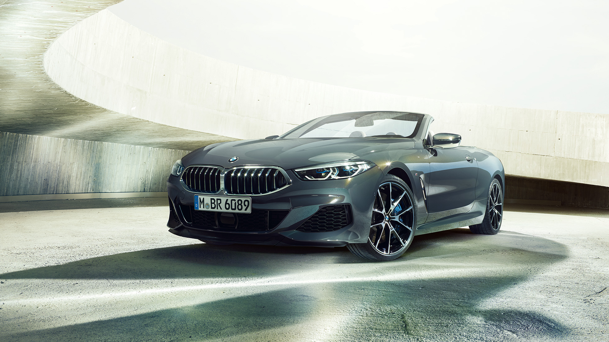 Imagine cu trei sferturi din partea frontală a BMW M850i xDrive Cabriolet. Imagine cu trei sferturi din partea frontală a BMW M850i xDrive Cabriolet în Gri Dravit metalizat cu soft-top-ul deschis, staţionând în faţa unei clădiri de beton.