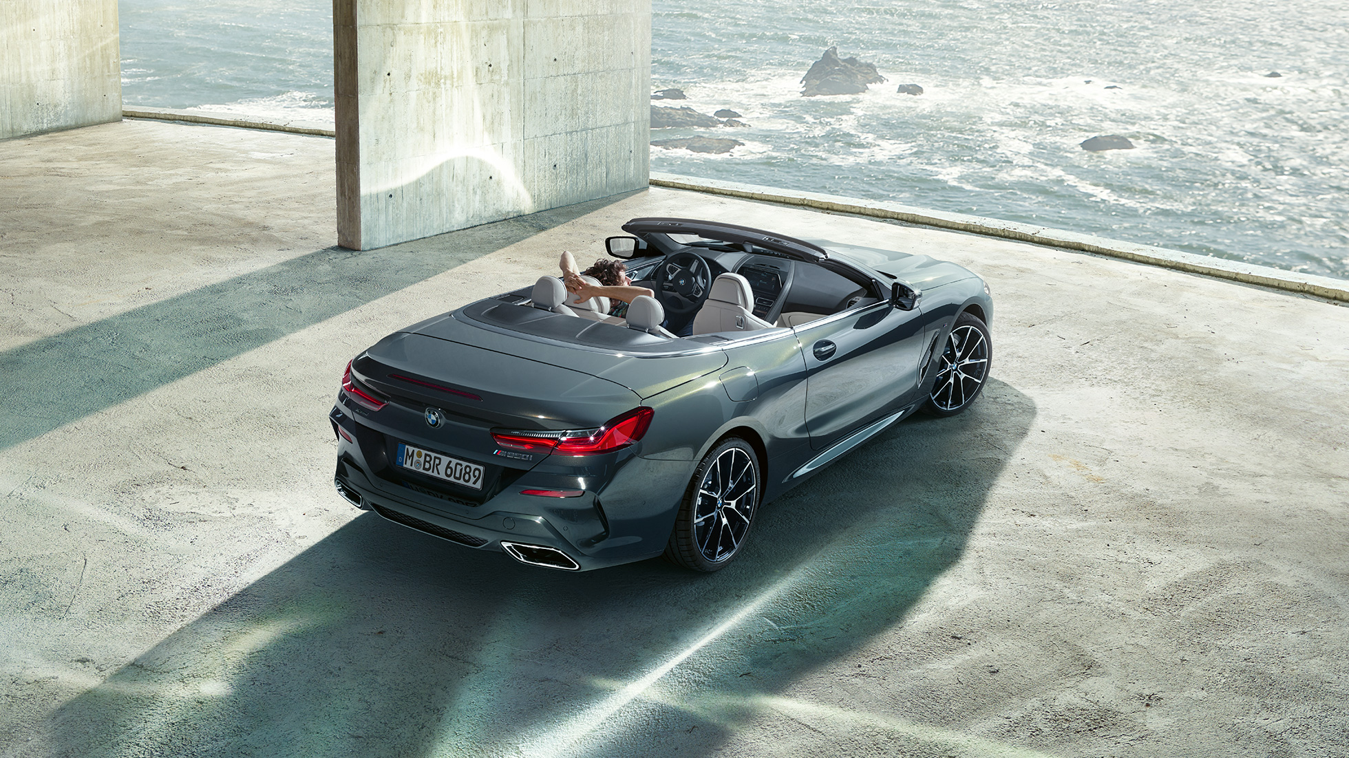Perspectivă de sus asupra BMW M850i xDrive Cabriolet. Perspectivă de sus asupra BMW M850i xDrive Cabriolet în Gri Dravit metalizat cu soft-top-ul deschis, parcând într-o parcare de beton în faţa oceanului.