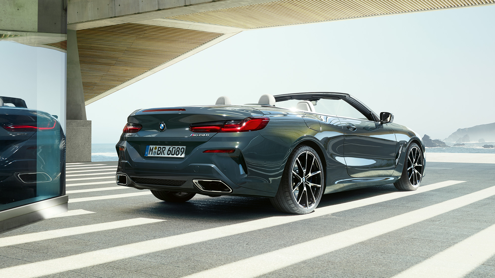 Imagine cu trei sferturi partea spate a BMW M850i xDrive Cabriolet. Imagine cu trei sferturi din partea spate a BMW M850i xDrive Cabriolet în Gri Dravit metalizat cu soft-top-ul deschis, parcând cu oceanul pe fundal.