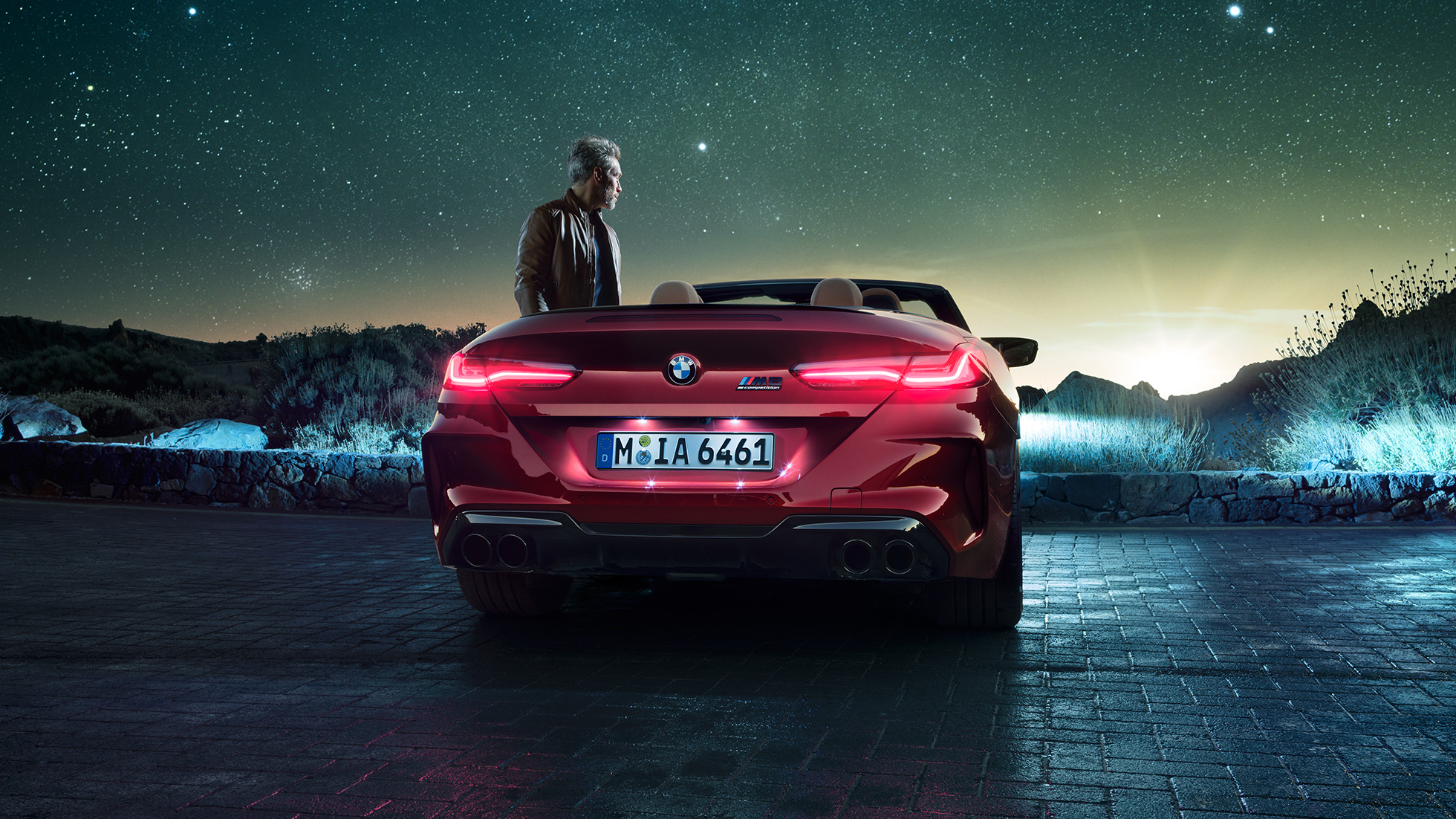 Imagine cu partea spate a BMW M8 Competition Cabriolet. Imagine cu partea spate a BMW M8 Competition Cabriolet în Roşu Motegi metalizat cu capota deschisă, staţionând alături de un bărbat sub cerul înstelat.