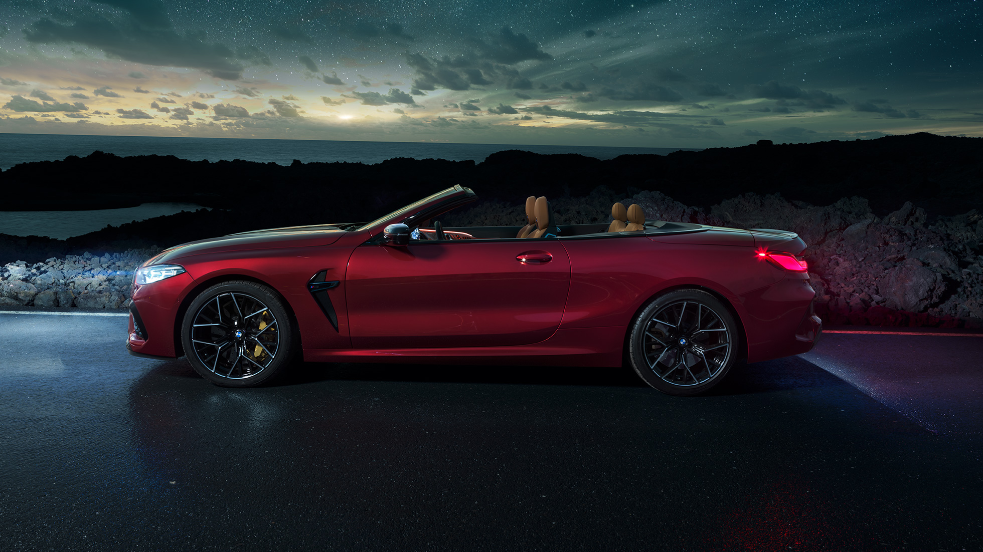 Imagine cu partea laterală a BMW M8 Competition Cabriolet. Imagine cu partea laterală a BMW M8 Competition Cabriolet în Roşu Motegi metalizat cu soft-top-ul deschis, staţionând noaptea sub cerul înstelat.