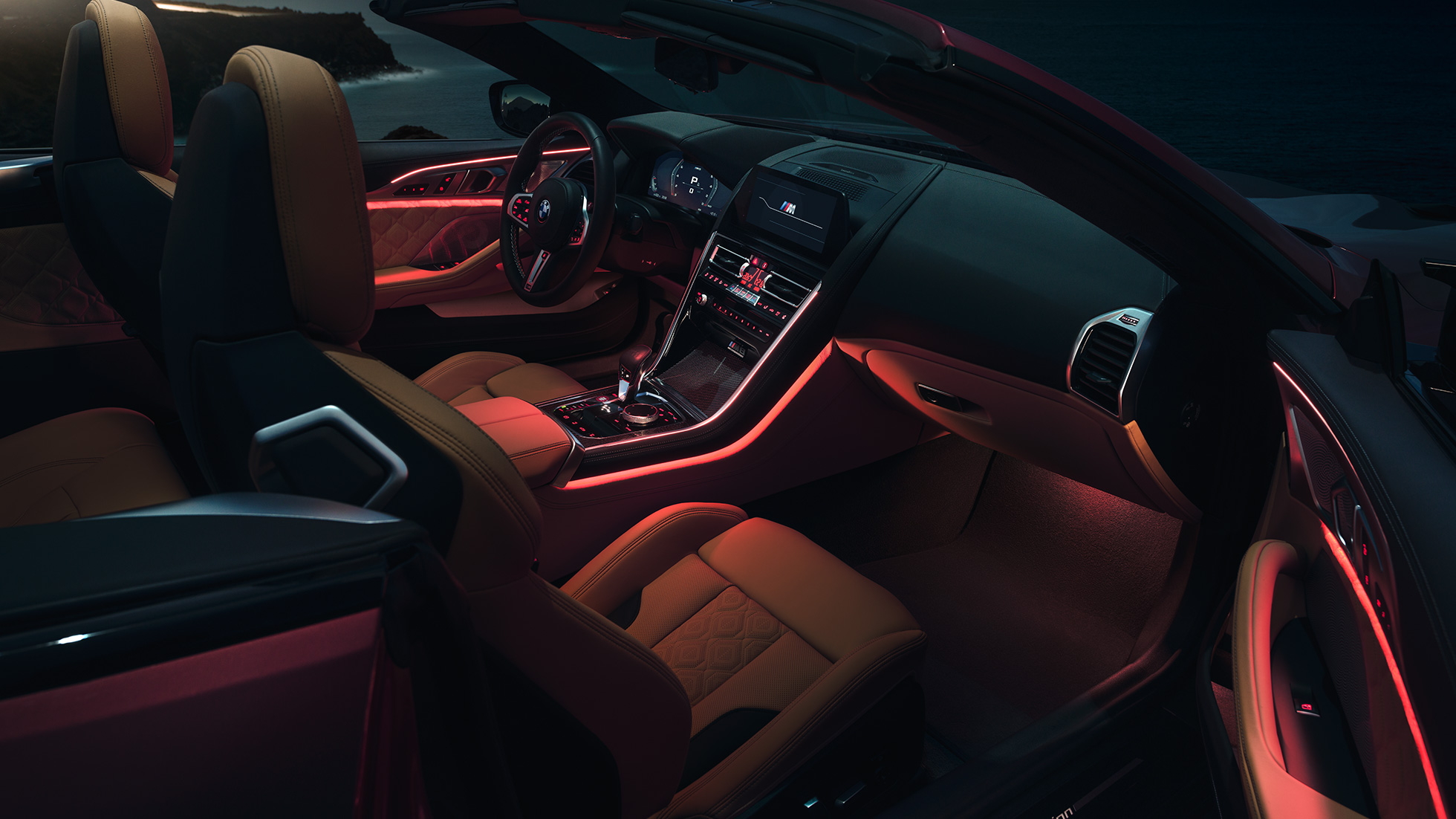 Interiorul BMW M8 Competition Cabriolet. BMW M8 Competition Cabriolet cu soft-top-ul deschis şi interior cu iluminare ambientală roşie, pe timp de noapte.