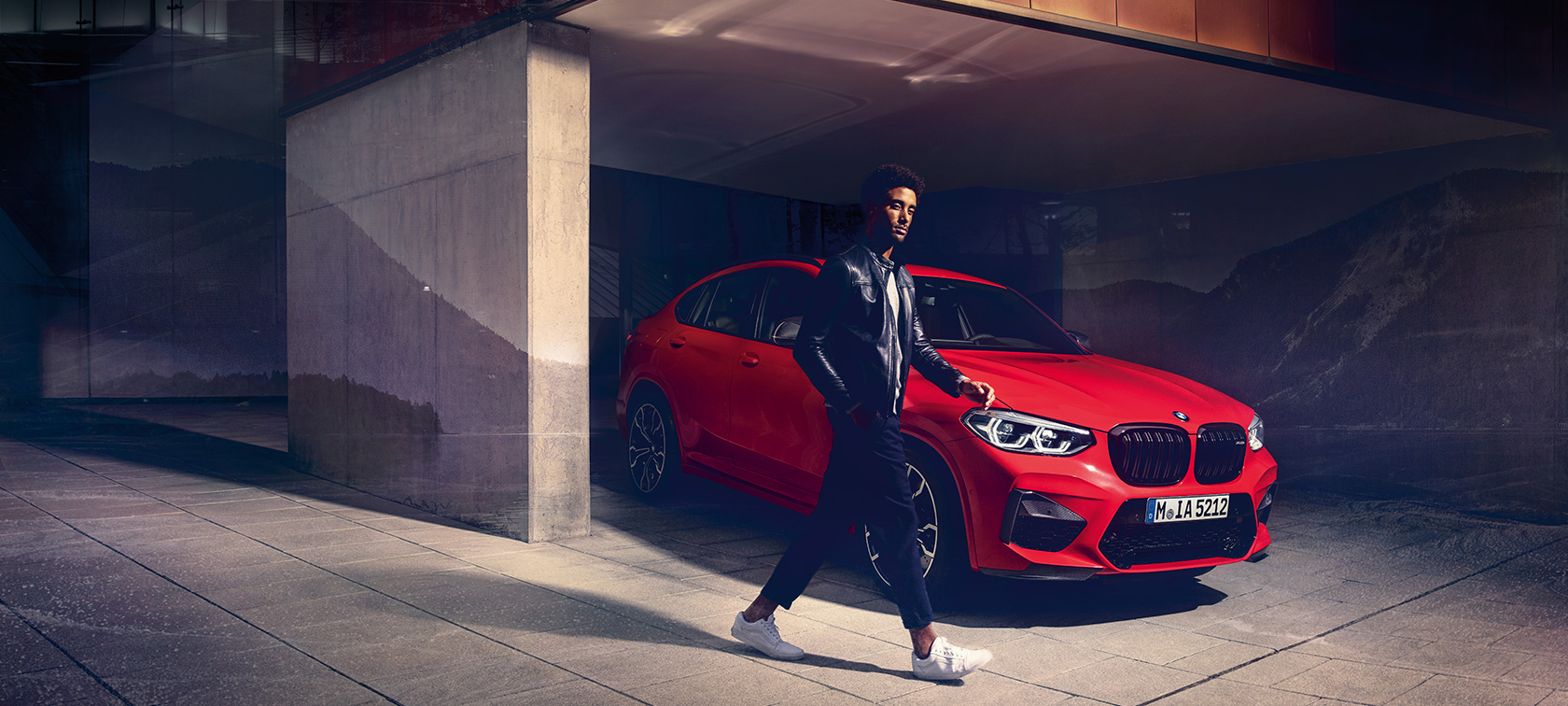 Imagine cu trei sferturi din partea laterală exterioară a BMW X4 M Competition în Roşu Toronto metalizat alături de un tânăr.