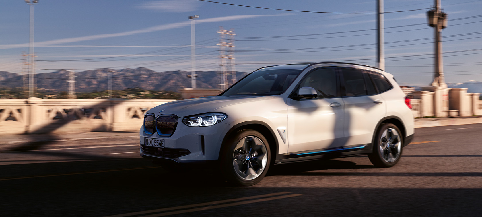 Imagine cu trei sferturi din partea frontală a BMW iX3, G08, rulând pe şosea Imagine cu trei sferturi din partea frontală a BMW iX3, G08, 2020, în Alb Mineral metalizat