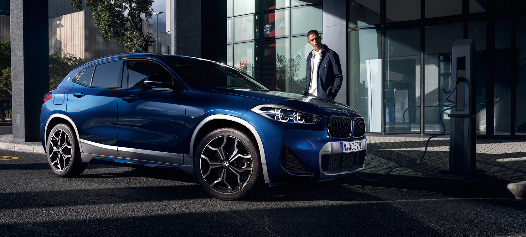 Imagine cu trei sferturi din partea laterală a BMW X2 xDrive25e F39 plug-in hybrid la o staţie de încărcare Imagine cu trei sferturi din partea laterală a BMW X2 xDrive25e F39 plug-in hybrid la o staţie de încărcare cu un model