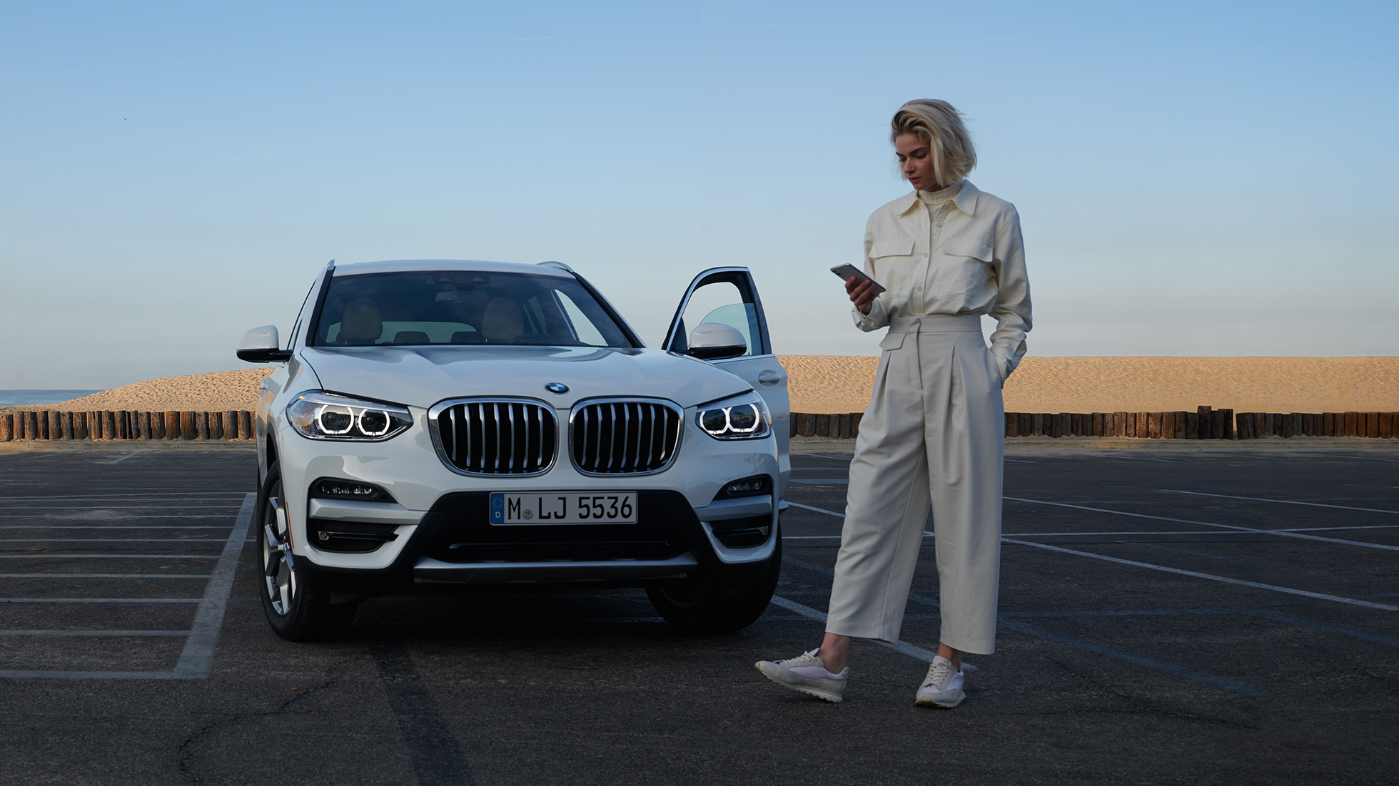 Femeie care utilizează aplicaţia My BMW pe smartphone Femeie utilizând aplicaţia My BMW din BMW ConnectedDrive pe smartphone în faţa unui BMW X5 alb