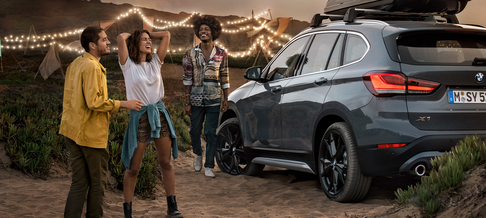 Un trio bucuros într-un automobil BMW X1, echipat cu accesorii BMW de vară Imagine cu un trio bucuros într-un automobil BMW X1 în Gri Mineral, echipat cu accesorii BMW de vară, staţionând la plajă, oferte de vară BMW