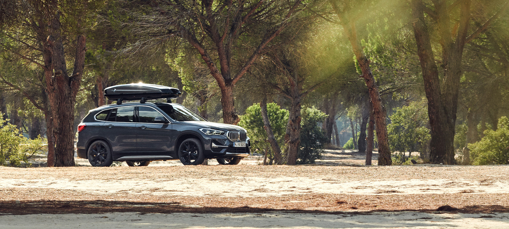 Imagine cu partea laterală a BMW X1, echipat cu accesorii de vară şi cu cutie BMW pentru plafon Imagine cu partea laterală a BMW X1 în Gri Mineral, echipat cu accesorii de vară şi cu cutie BMW pentru plafon, staţionând într-o pădure
