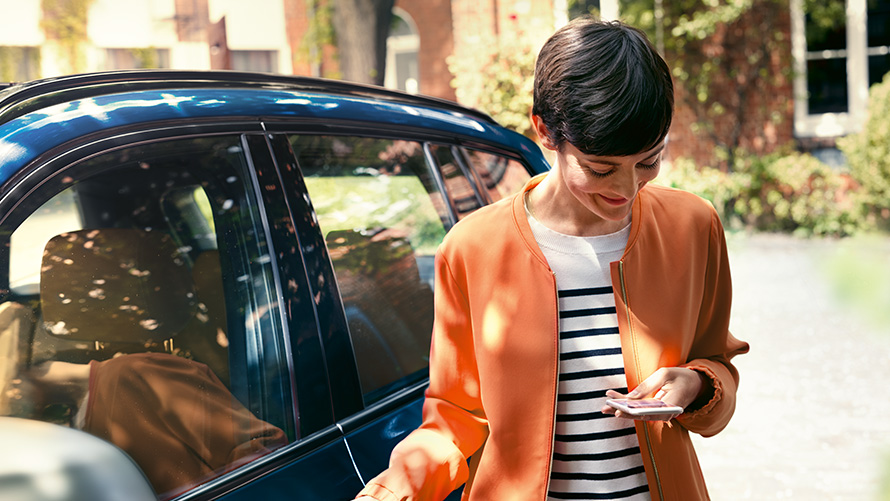 BMW Teleservices, oferte de vară BMW Imagine cu o femeie care stă în faţa unui automobil BMW şi foloseşte un smartphone, BMW Teleservices, oferte de vară BMW