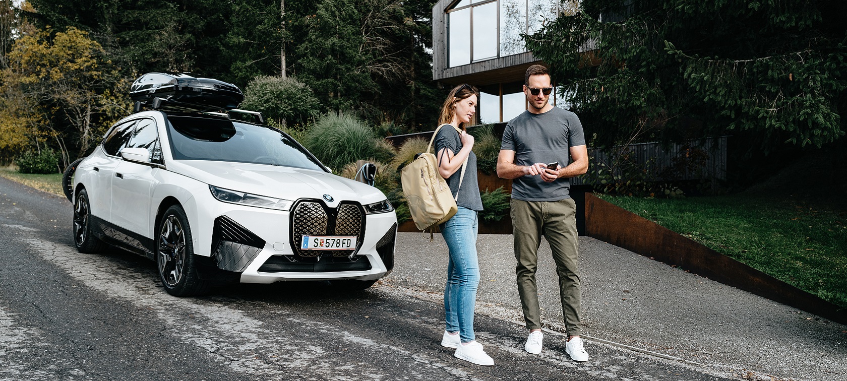 Man with phone and woman with backbag standing in front of a BMW iX. Man with phone and woman with backbag standing in front of a BMW iX.